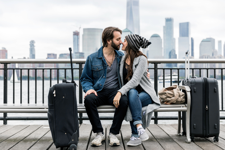 Young Couple Travelers Sitting on Bench and Waiting with Suitcases, bag with Lower Manhattan and the World Trade Center in the background.