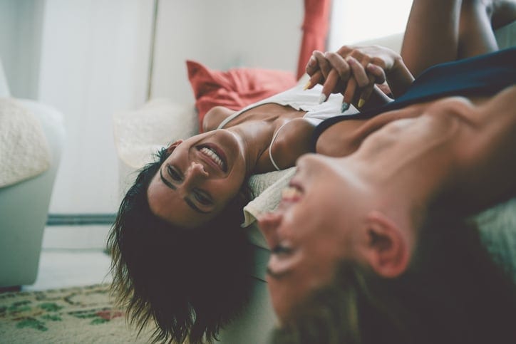 Two Laughing Sensual Females Lying on Sofa and Holding Hands Upside Down