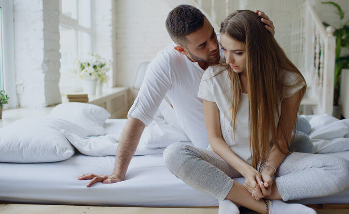 Depressed young woman sitting in bed and crying while her boylfriend come and embrace her and kiss in bedroom at home