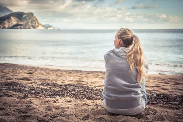 Pensive lonely young woman tourist sitting on beach hugging her knees and looking into the distance with hope