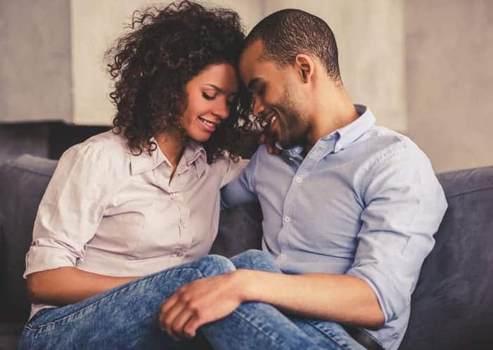 Beautiful young Afro American couple is hugging and smiling while sitting together on couch at home