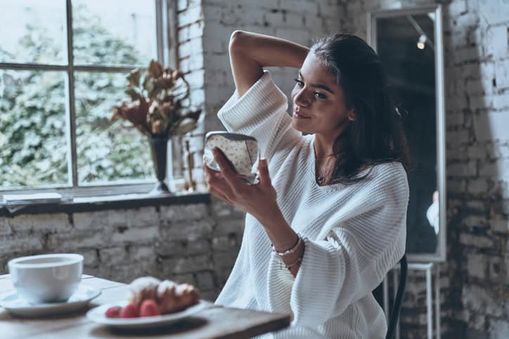 Young woman sitting at breakfast table, looking at herself in pocket mirror