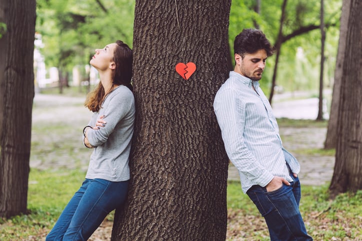 Unhappy couple leaning on opposite sides of tree trunk.