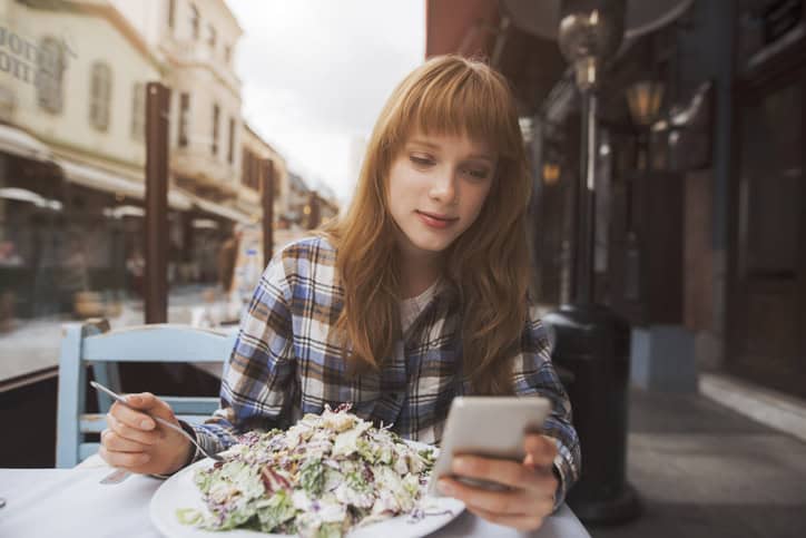 beautiful young woman is eating a salad in a restaurant while using an application in her smart phone
