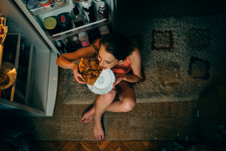 Woman sitting and eating a snack at the refrigerator.