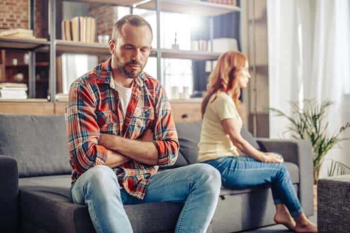 Upset husband turning away from his wife after quarrel while sitting on sofa at home