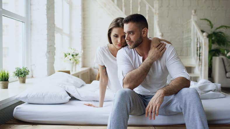Depressed yong man sitting in bed having stressed while his girlfriend come and embrace him in bedroom at home