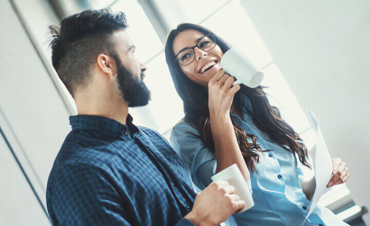 Closeup of two late 20's people taking a coffee break at their office. They are standing next to a window and having casual funny conversation. Tilt shot.