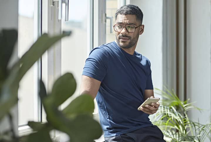 Mixed race male weekend worker looking out the window with concerned expression after learning bad news on smart phone.