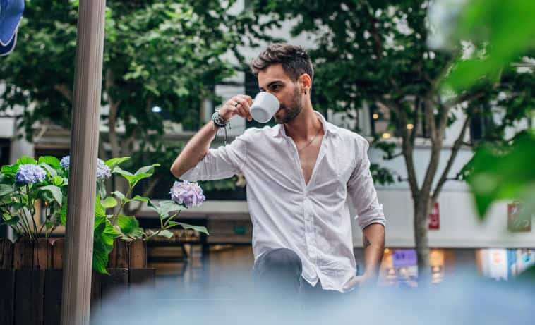 Handsome modern man with white shirt and beard, standing outdoors and drinking coffee.