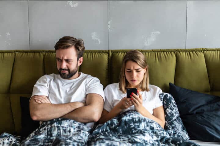 Confused young couple having an argument about mobile phone while lying in bed
