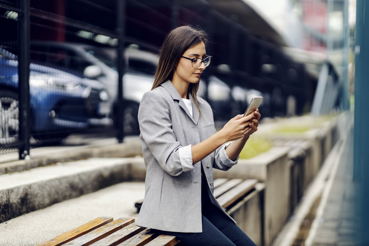A successful young businesswoman dressed smart casual is sitting outdoors and replying messages to a satisfied client. Nowadays, everywhere is our workplace.