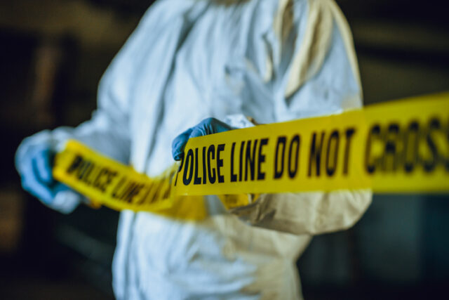 Forensic detective with crime tape at a scene.