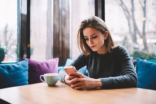 Concentrated young female teenager sitting at cafe and reading messages on mobile phone while connected to 4g internet, serious woman spending time with modern smartphone and morning cup of coffee