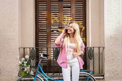 Trendy young blonde woman stop to riding on her vintage bike with basket of flowers while focused chatting or talk on smart phone outside. High quality photo