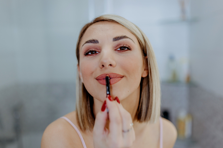 Woman applying lipstick in bathroom mirror.