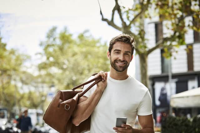Portrait of smiling businessman carrying bag and holding smart phone. Confident professional is wearing white t-shirt. He is in city.