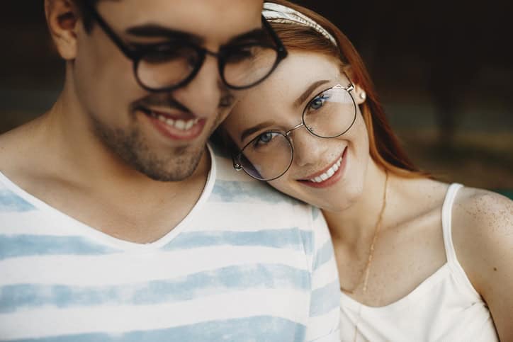 Close up portrait of a young couple sitting closely while smiling. Red haired girl with freckles looking at camera while leaning head on her boyfriend.