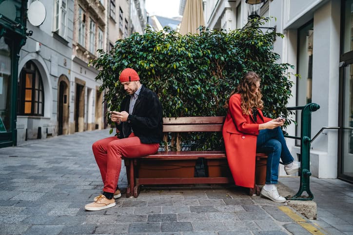Young Caucasian man and woman sitting back to back on bench and using phones, not looking to each other.