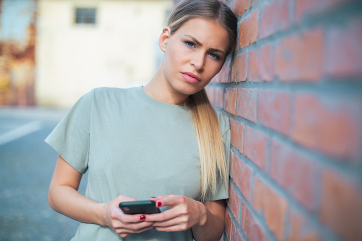 Frustrated woman texting by brick wall.