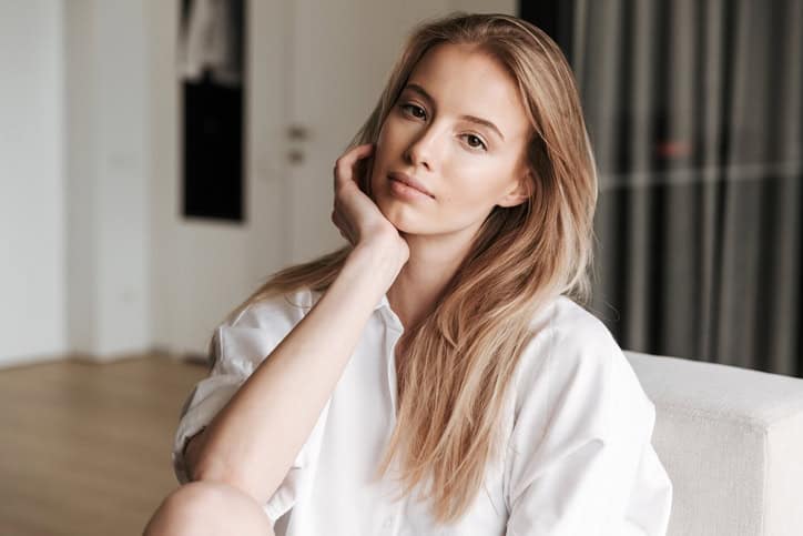 Beautiful young woman dressed in white shirt sitting on a couch at home and looking at camera