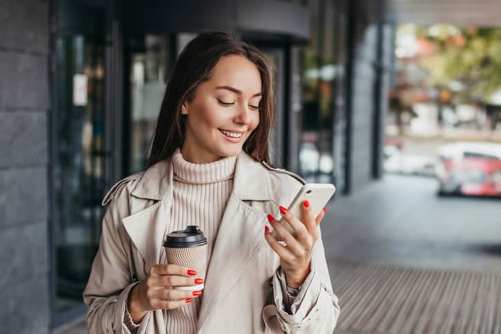 Young smiling ukrainian brunette woman holding a smartphone and a cup of coffee looking at device screen against the background of an office building. copy space