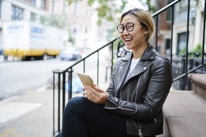 Cheerful Asian girl laughing at joke from social networks while sitting on stairs, happy Chinese female in glasses using application on smartphone for browsing phone mobility spending time in downtown