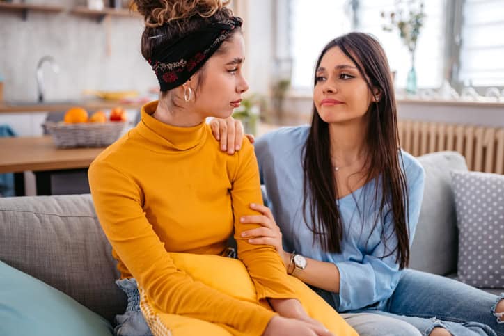 A caring young woman sitting on a sofa in the living room and comforting her sad friend that is having problems.