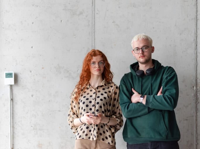 ir standing together, their arms crossed, posing confidently in front of a gray wall, exuding a sense of style and professionalism.