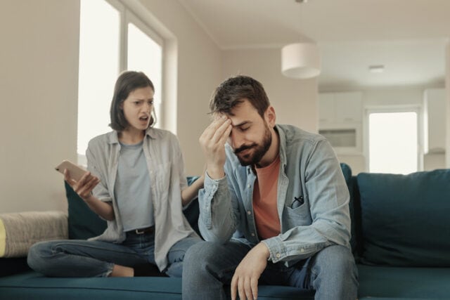 Angry couple or marriage fighting for a mobile phone at home. Jealous caucasian woman holding smart phone and showing message to his husband. Angry girlfriend asking for an explanation to her cheater boyfriend, point at his smartphone
