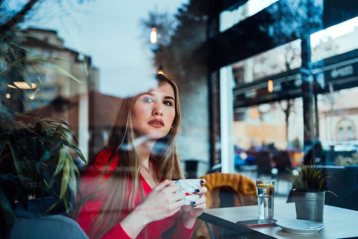 Young caucasian woman drinking coffee in cafe, looking through the window and waits for a friend