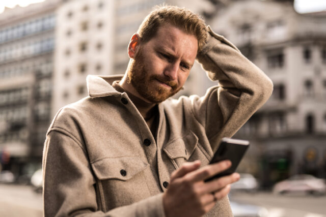 Frustrated and distraught young man reading something on his phone on city street.