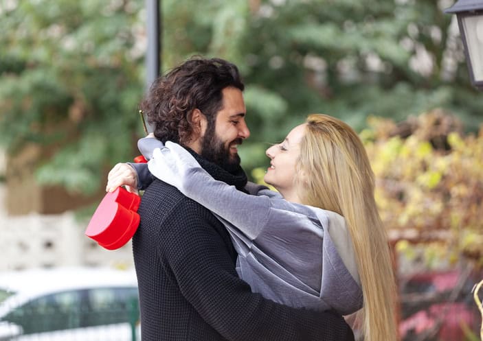 Young man hugging his girlfriend from behind and surprising her with a red rose.