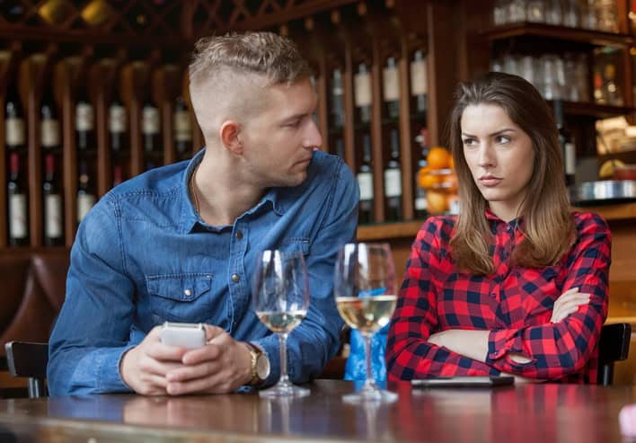 man holding phone, text messaging, sulking woman is looking him in restaurant