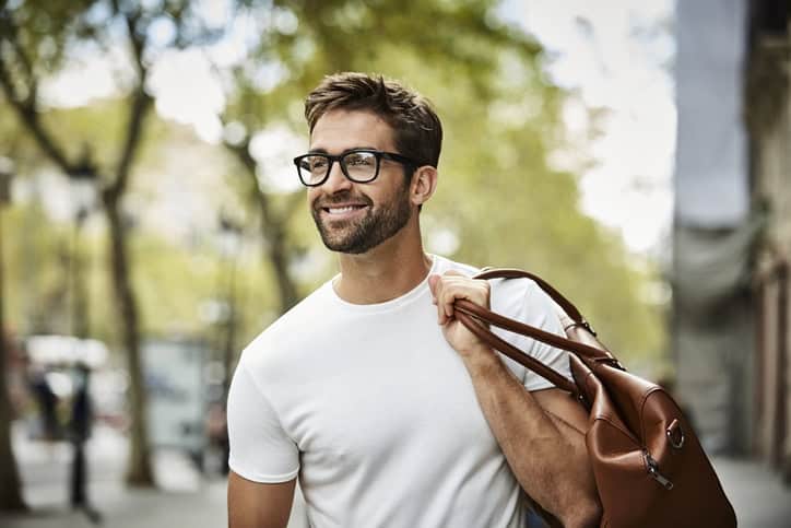 Smiling handsome businessman with beard walking in city. Executive is looking away while carrying bag. He is wearing eyeglasses.