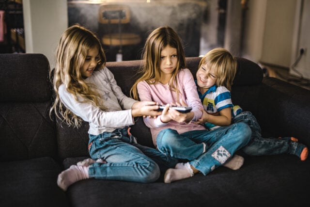 Angry little kids fighting over a remote control while watching TV on sofa at home.