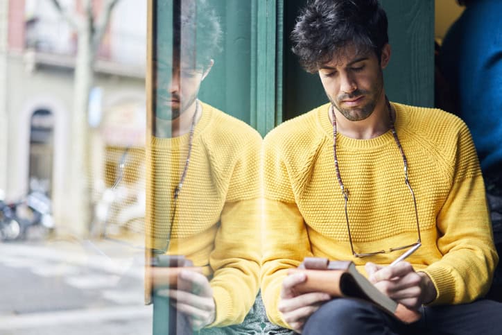 Crop concentrated bearded man in casual clothes writing in notebook while sitting near big window in street cafe during daytime