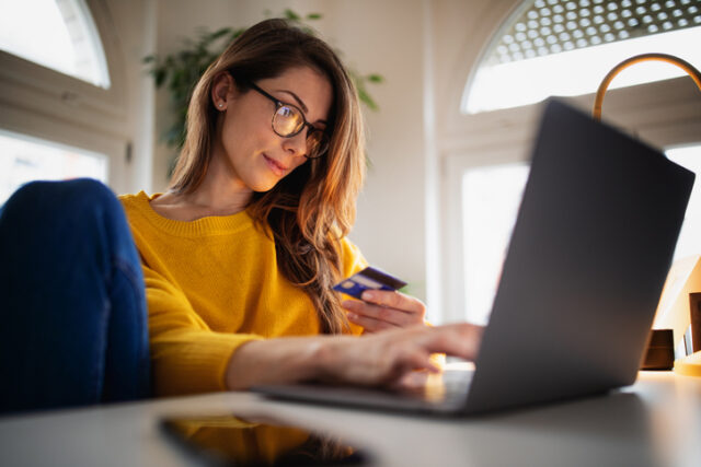 A beautiful young woman enjoying working at home on her laptop in cozy and bright apartment wearing yellow sweater and shopping online paying with credit card
