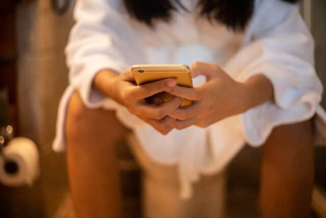 Woman holding a phone while using the toilet.