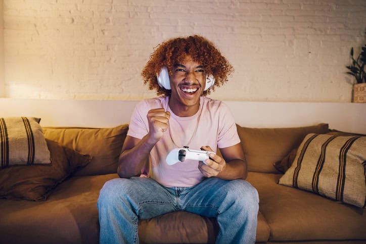 Young man with white headphones playing video games on a sofa in a living room. Happy young man celebrating a winning