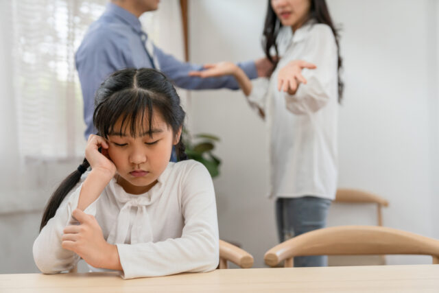 Sad young girl sitting alone while her parents are arguing behind her.