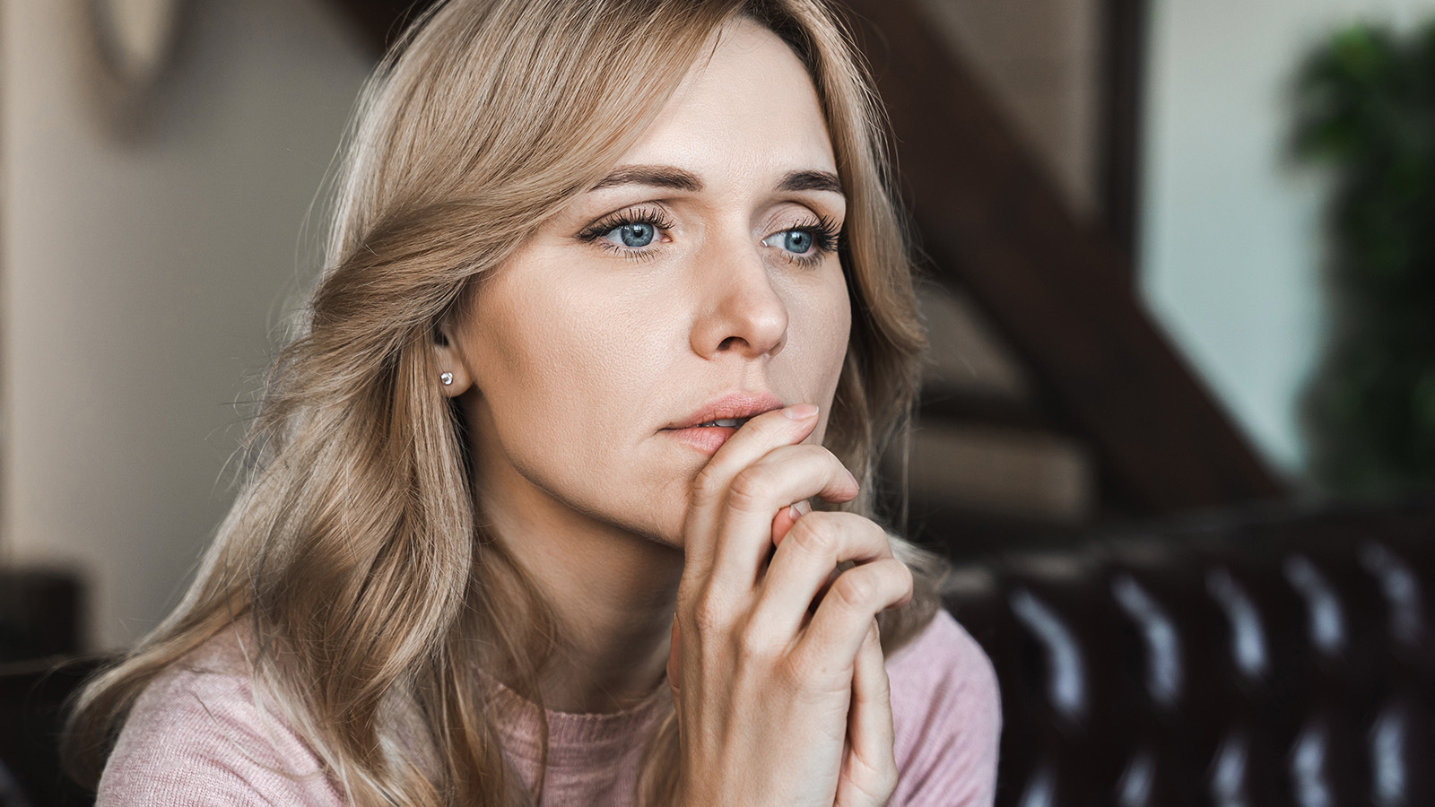A woman with long blonde hair sits indoors, resting her chin on her hands and looking thoughtfully into the distance, her expression conveying a sense of loneliness that feels deeply relatable.