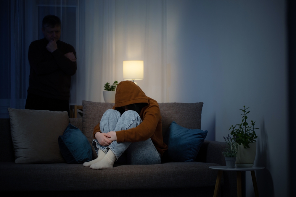 A teenage girl in the living room at night, wearing a hoodie and jeans and resting her head on her knees, her father us in the shadows staring at her