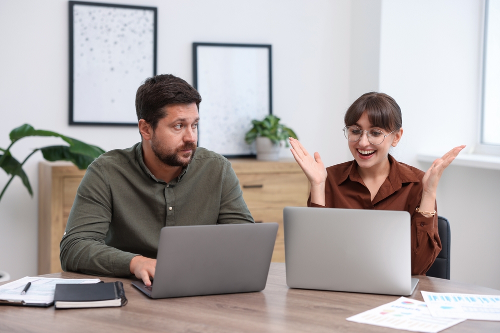 Two colleagues working with their laptop at work and the man looks jealous of the woman.