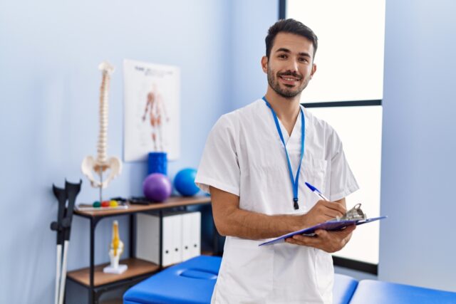 Male physiotherapist in a consultation room, holding a clipboard.