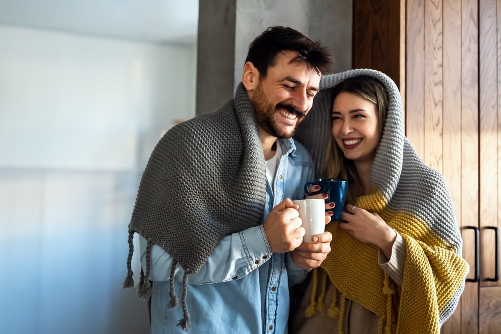 Happy couple keeping warm under a blanket drinking hot tea.