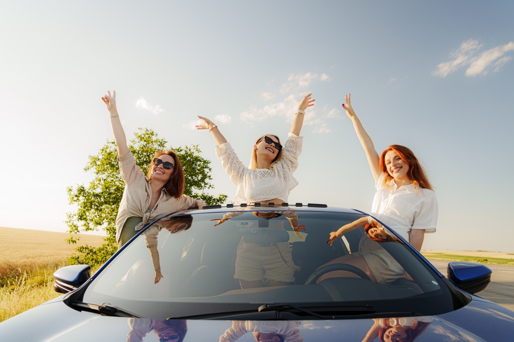 Three female friends popping out of a moving car sunroof with their arms raised in the air