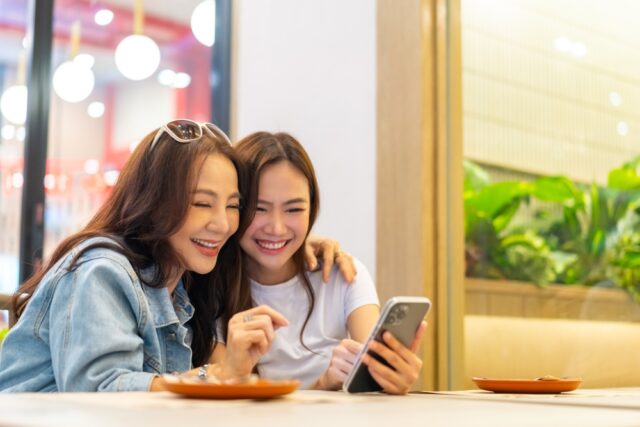 Happy Asian mother and daughter using smartphone together, having lunch eating food together in restaurant.
