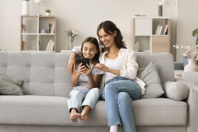 Mother and young daughter sitting on sofa, looking at phone smiling.
