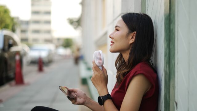 Young woman sitting down holding handheld fan to her face.